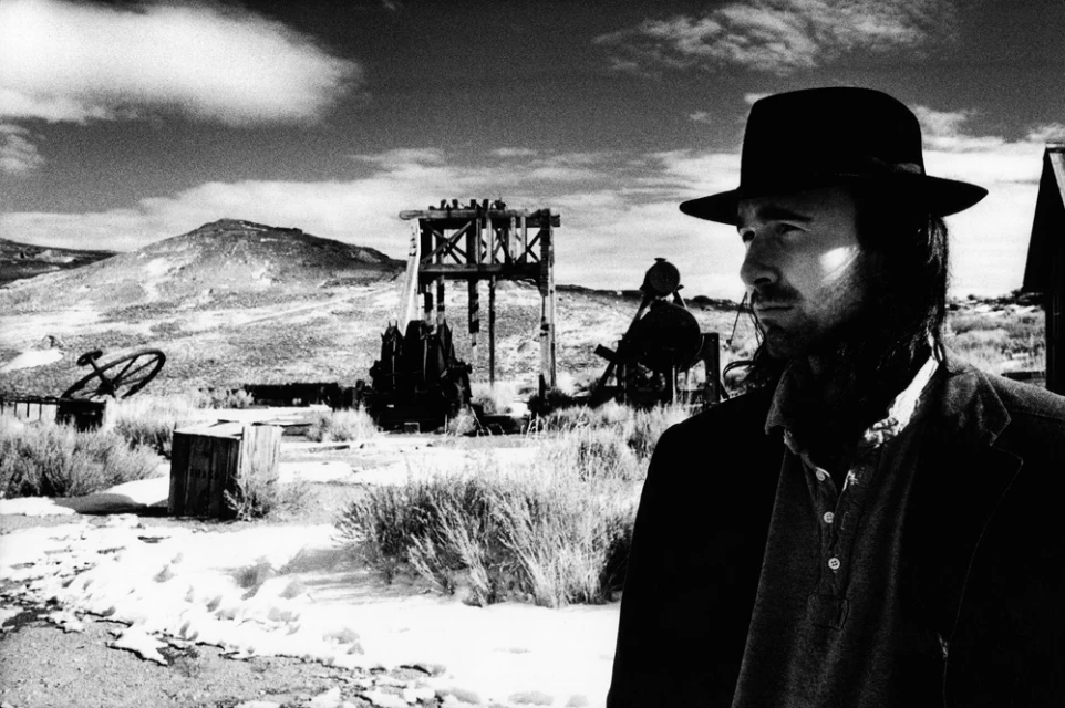 Black and white outdoor portrait of a man in a hat with a Western-style set, featuring a construction and mountainous landscape.