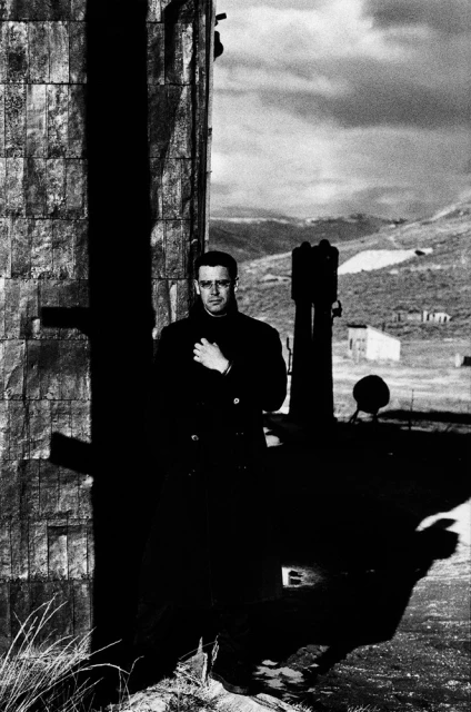 Monochrome portrait of a man in a coat standing next to a textured wall against a cloudy sky and hilly landscape. 