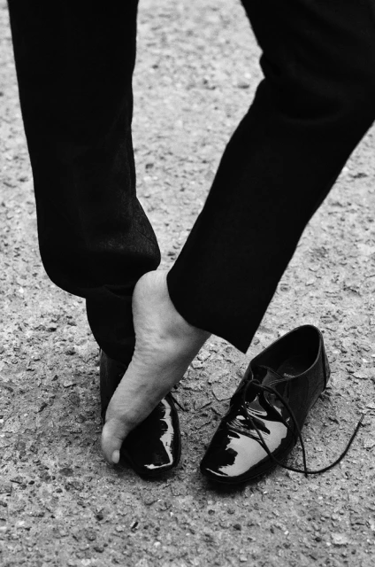 A person, seen from the knees down, removes a shiny, black dress shoe on a textured surface in a black-and-white, high-contrast photo.