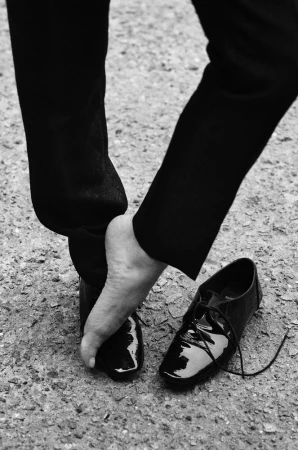 A person, seen from the knees down, removes a shiny, black dress shoe on a textured surface in a black-and-white, high-contrast photo.