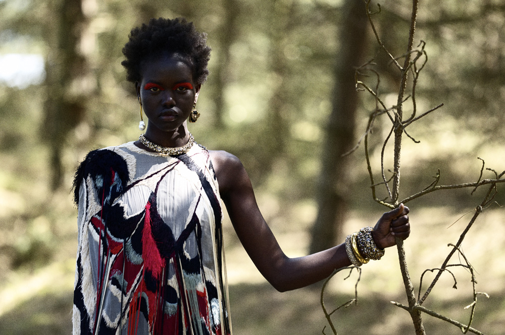Woman with red eye makeup and feathered dress poses in a forest setting, accessorized with a statement necklace and cuff bracelets.