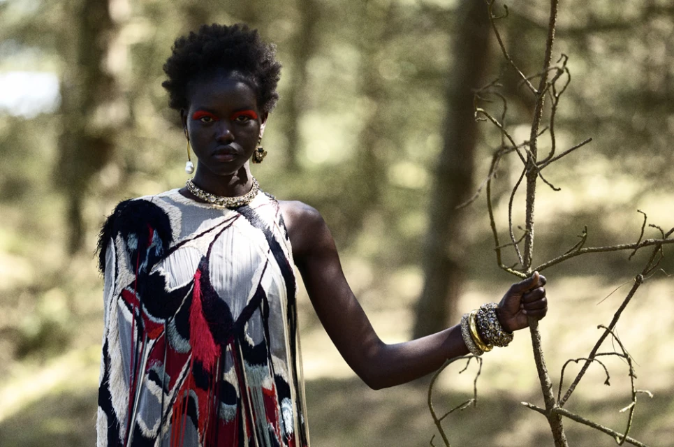 Woman with red eye makeup and feathered dress poses in a forest setting, accessorized with a statement necklace and cuff bracelets.