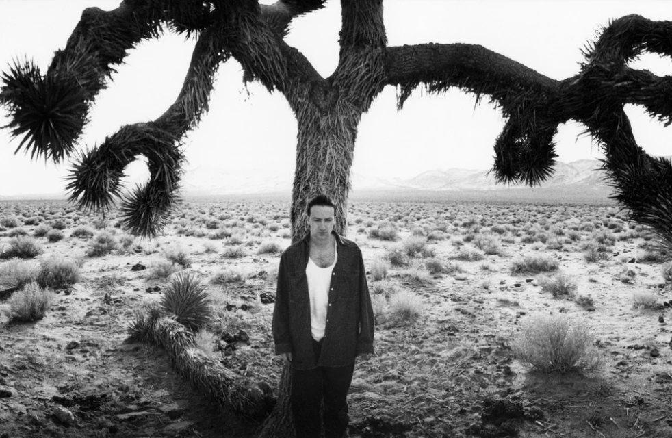 Black and white portrait of a man in front of a large Joshua tree in a desert landscape, with low mountains on the horizon.