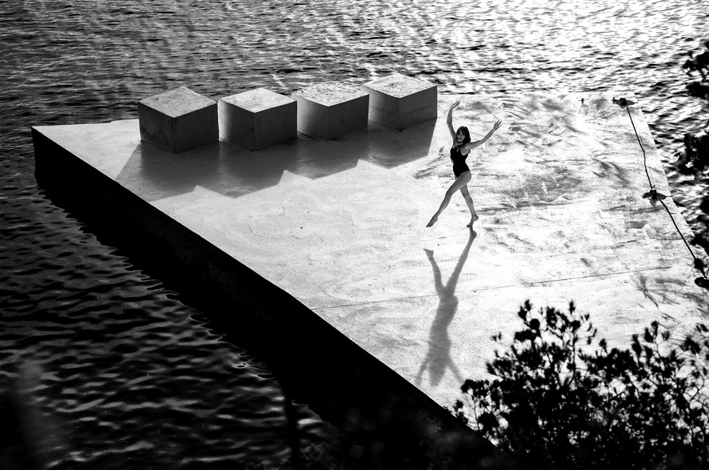 Black and white photo of a woman in a swimsuit dancing on a concrete platform with cube props surrounded by dark water and foliage.