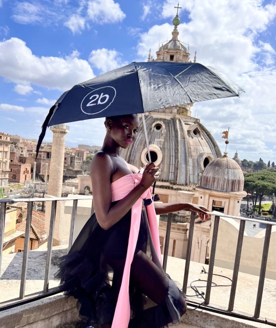 Fashion shoot on rooftop overlooking Roman cityscape featuring a Black female model in a pink and black gown holding an umbrella.