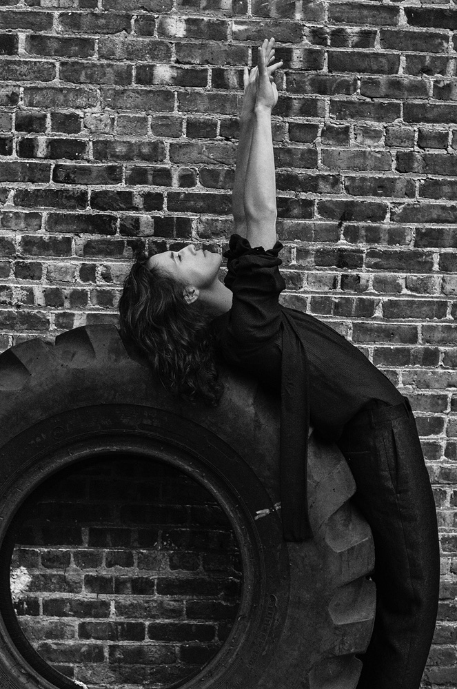 Monochrome portrait of a woman in dark clothing leaning back on a large tire against a brick wall, arms raised, looking upward.