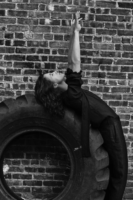 Monochrome portrait of a woman in dark clothing leaning back on a large tire against a brick wall, arms raised, looking upward.