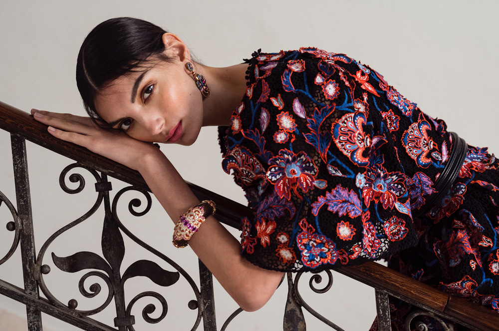 A woman with dark hair leans against an iron railing wearing a floral dress, earrings, and a bracelet in a close-up, eye-level fashion shot.