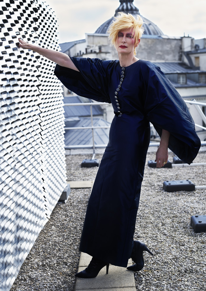 Woman with blonde hair and long black dress poses on a rooftop with architectural elements in the background on a cloudy day.