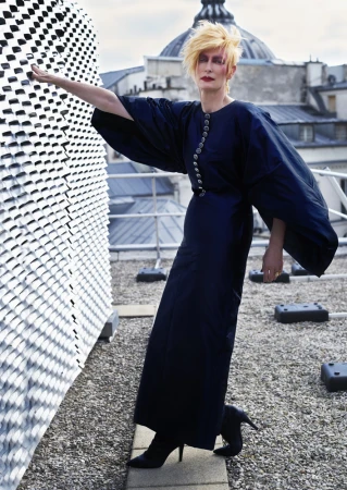 Woman with blonde hair and long black dress poses on a rooftop with architectural elements in the background on a cloudy day.