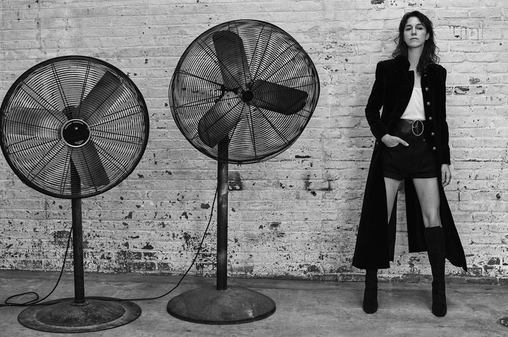Black-and-white shot of a woman in a dark coat and shorts, standing between two large industrial fans against a brick wall backdrop.