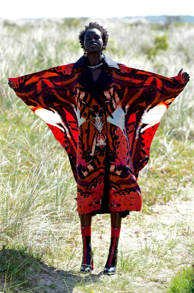A woman in a patterned red butterfly-wing coat stands in a grassy field. Fashion shot in natural light. Visually striking outdoor editorial.