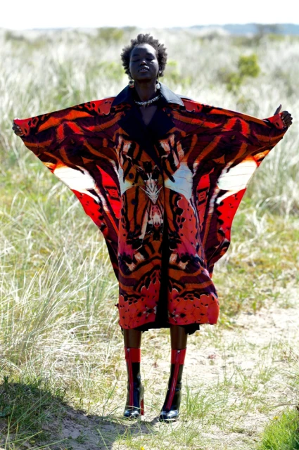 A woman in a patterned red butterfly-wing coat stands in a grassy field. Fashion shot in natural light. Visually striking outdoor editorial.