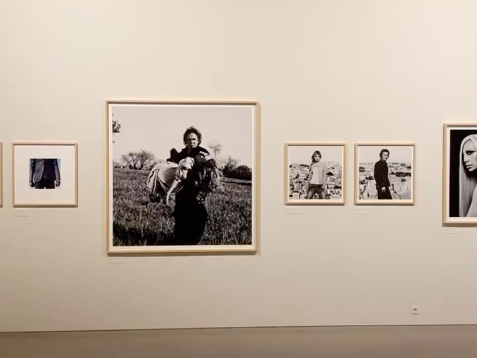 Monochrome photographs on display in an art gallery feature portraits of figures and couples against a plain wall in eye-level medium shots.