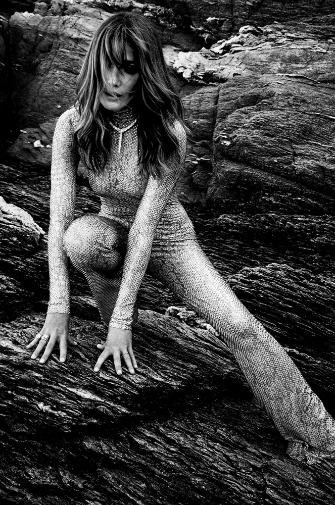 Woman in a long-sleeved jumpsuit poses on a rocky surface. The image is shot in black and white with natural lighting.