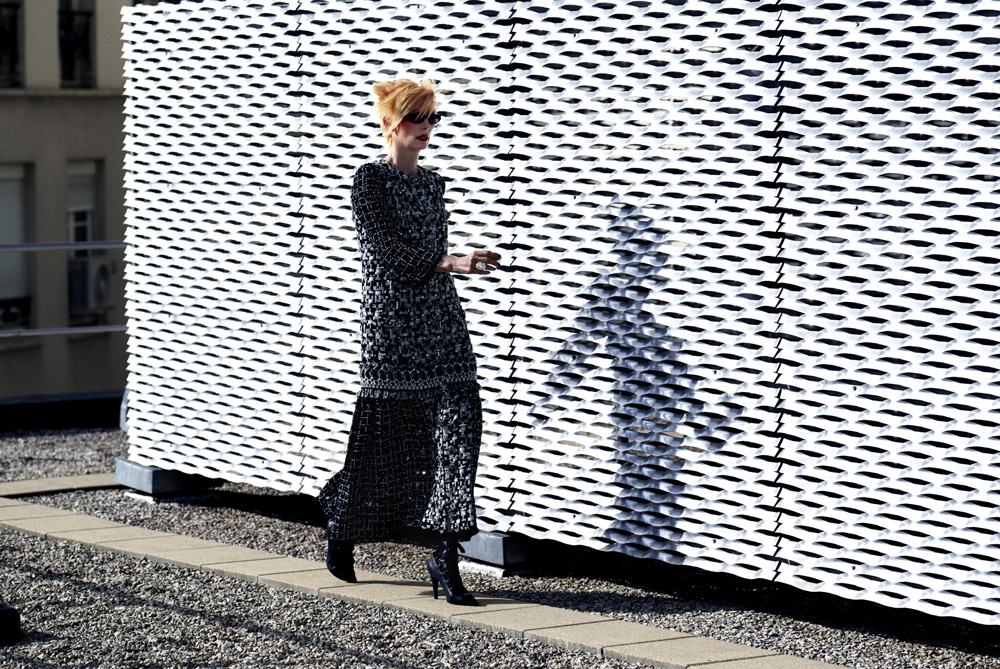 Adult woman in a black dress and sunglasses walks on a stone surface beside a white wall with a patterned design, on a sunny day.