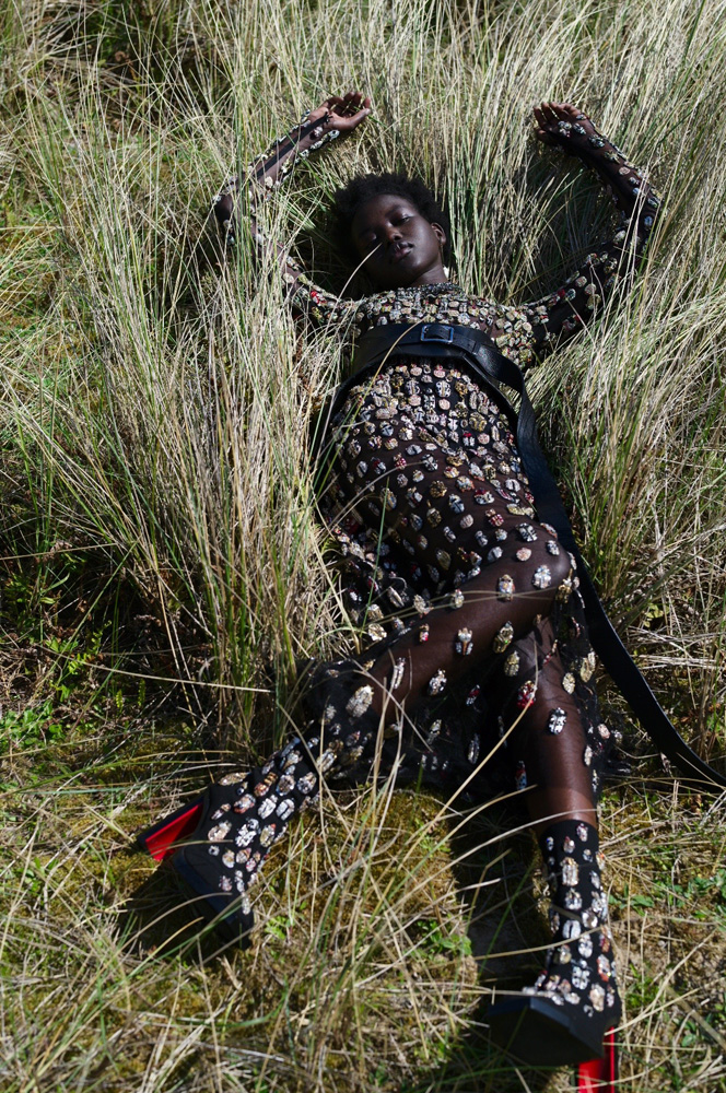 A woman lies in tall grass wearing a full-length black patterned dress, black belt, and black heels with red accents for a fashion shot.