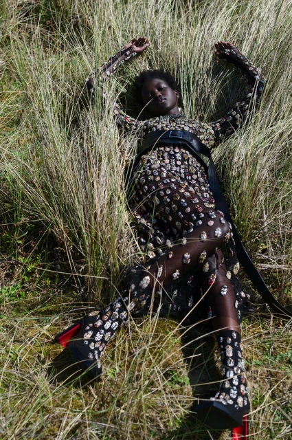 A woman lies in tall grass wearing a full-length black patterned dress, black belt, and black heels with red accents for a fashion shot.