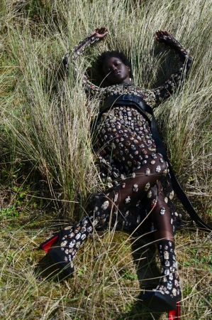 A woman lies in tall grass wearing a full-length black patterned dress, black belt, and black heels with red accents for a fashion shot.
