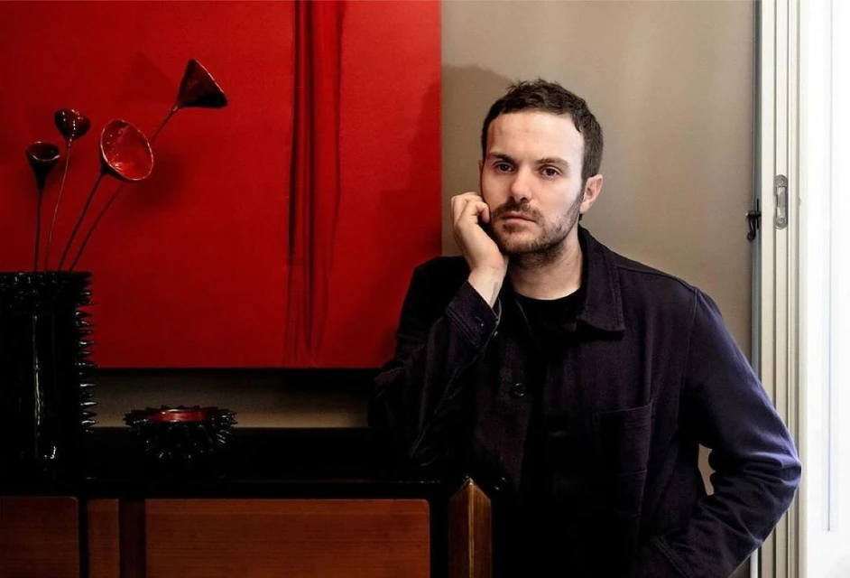 A portrait shot of a man indoors with a stoic expression, featuring a black vase with red and black metallic flower accents behind him.
