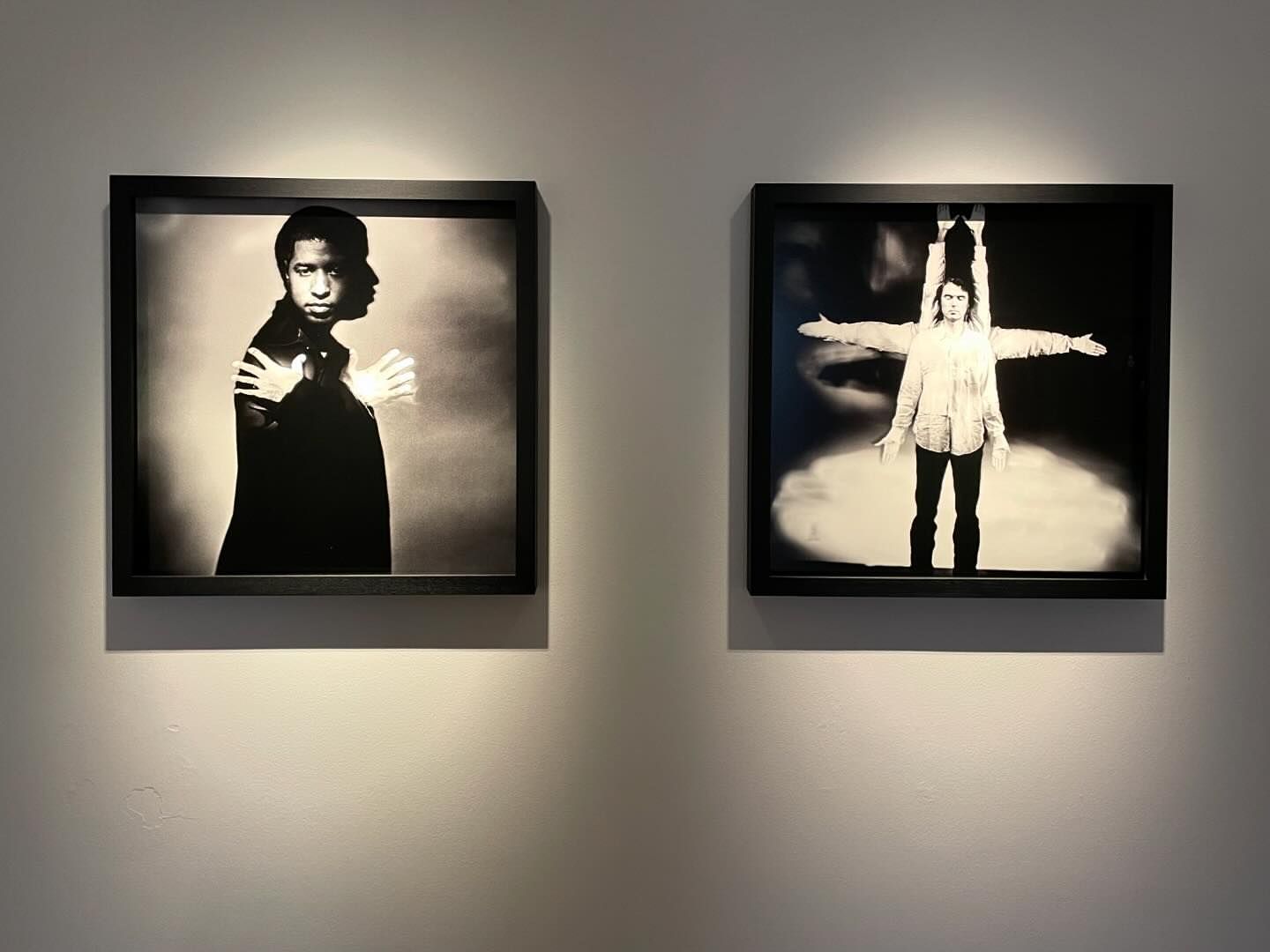 Two framed black and white photographs of people on display against a neutral wall, lit from above in an art gallery setting.