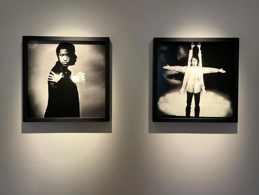 Two framed black and white photographs of people on display against a neutral wall, lit from above in an art gallery setting.