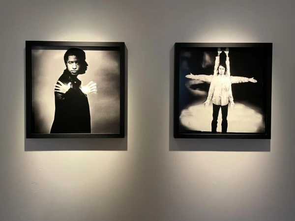 Two framed black and white photographs of people on display against a neutral wall, lit from above in an art gallery setting.