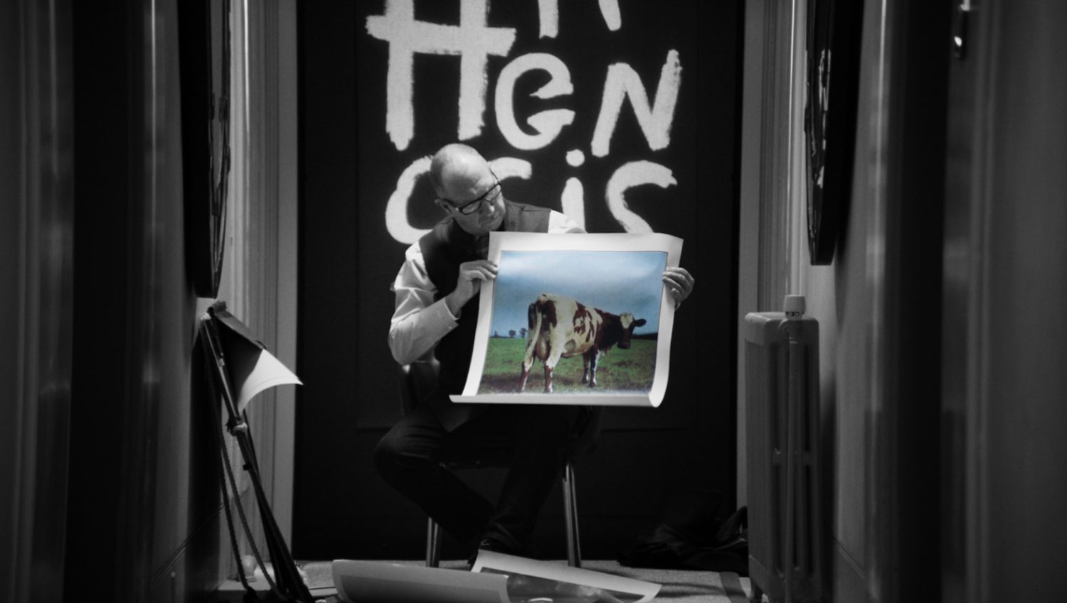 Man sits holding a picture of a cow in a field, in a black and white room with a heater and stage lighting. 