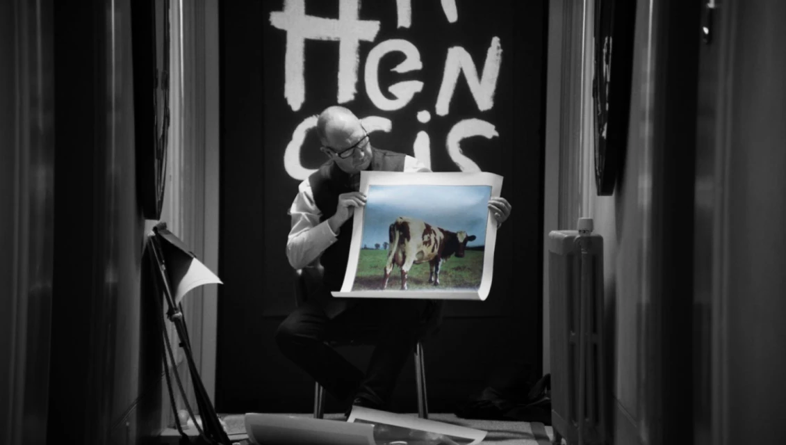 Man sits holding a picture of a cow in a field, in a black and white room with a heater and stage lighting. 