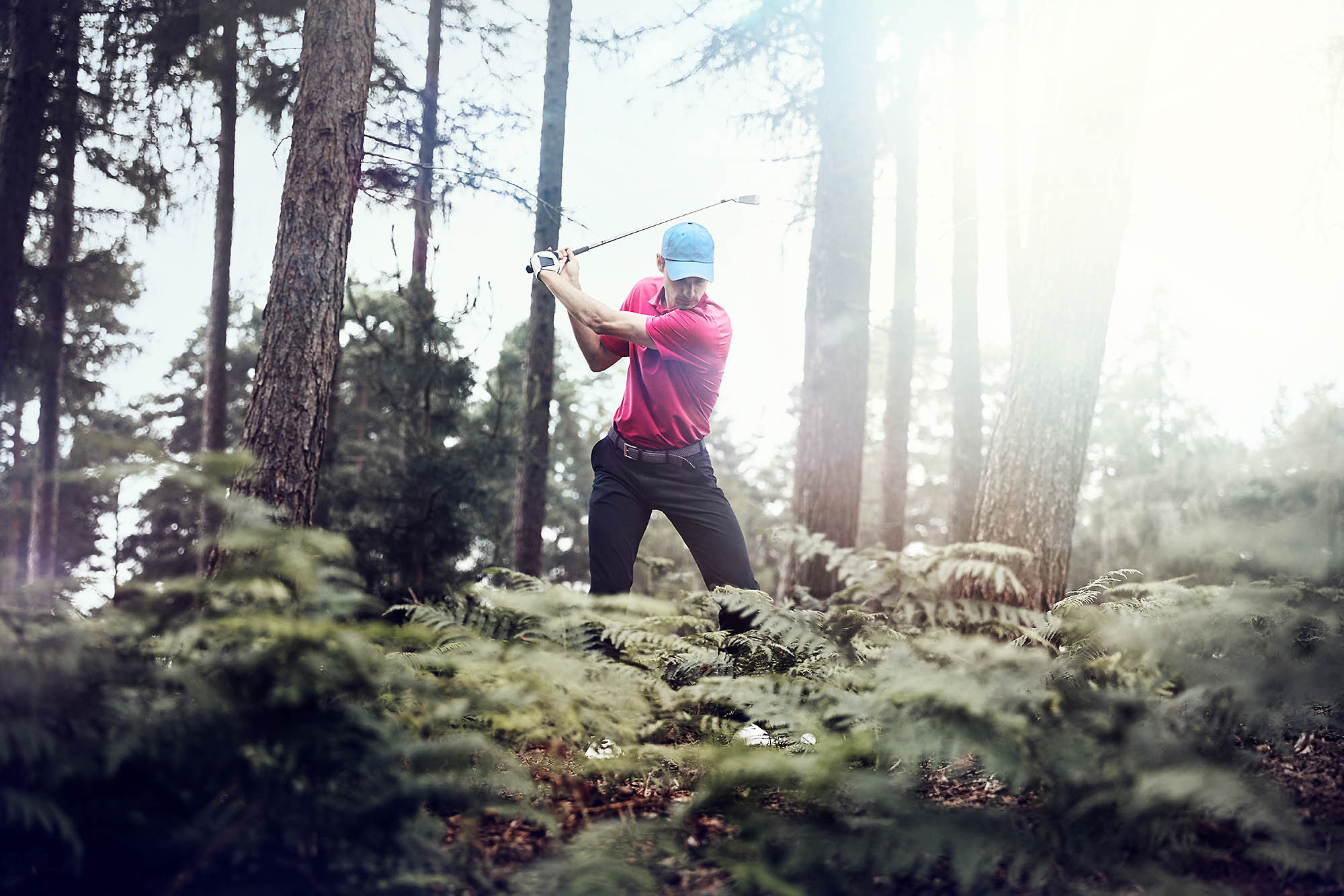 Man swinging golf club in forest. He's wearing a red shirt, black pants, and blue hat, surrounded by ferns and tall trees.