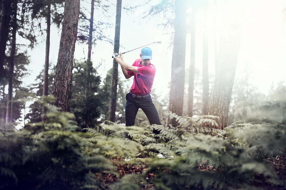 Man swinging golf club in forest. He's wearing a red shirt, black pants, and blue hat, surrounded by ferns and tall trees.