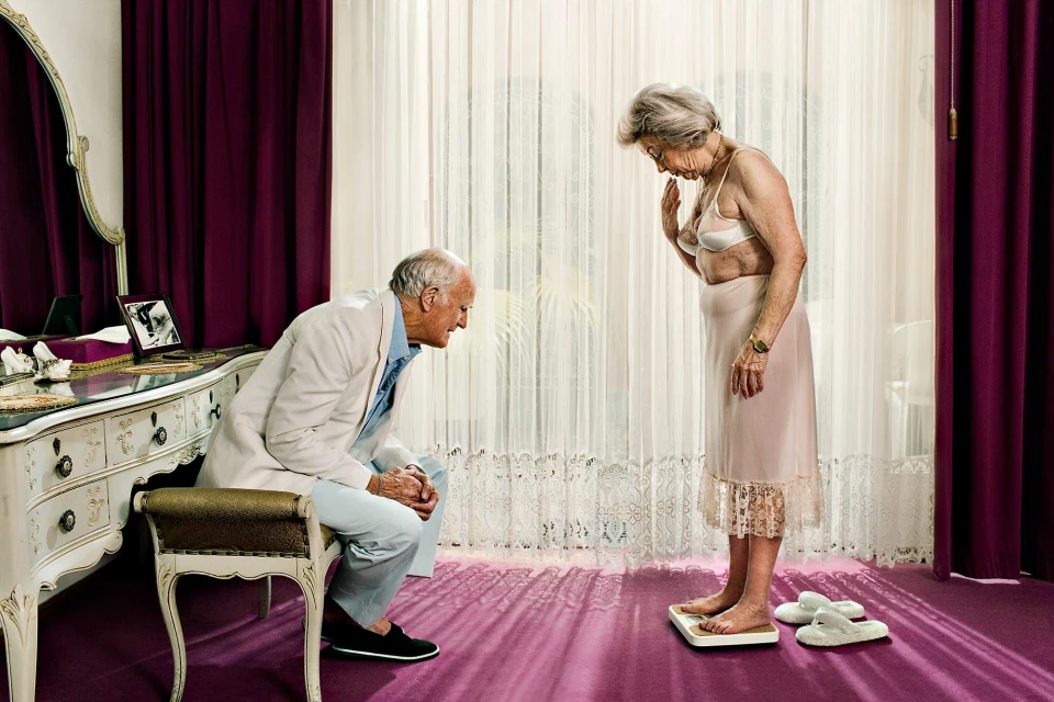 Elderly woman in bra and slip stands on scale as elderly man watches indoors, lit by window light and with purple carpet.