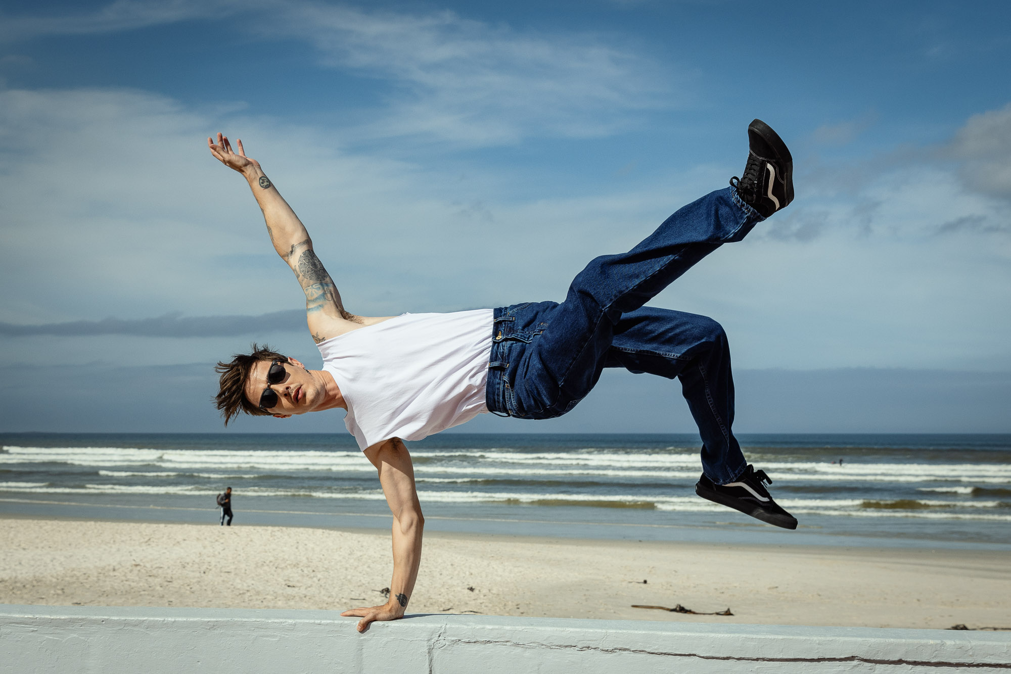 A man in mid-air, demonstrating a breakdancing move on a beach with blue sky and ocean waves in the background on a sunny day.
