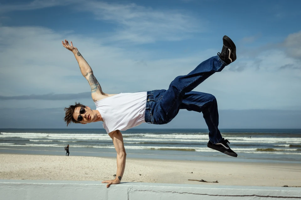 A man in mid-air, demonstrating a breakdancing move on a beach with blue sky and ocean waves in the background on a sunny day.