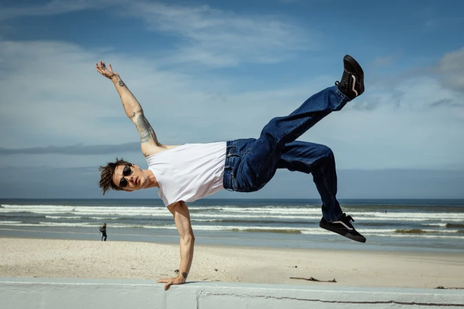 A man in mid-air, demonstrating a breakdancing move on a beach with blue sky and ocean waves in the background on a sunny day.