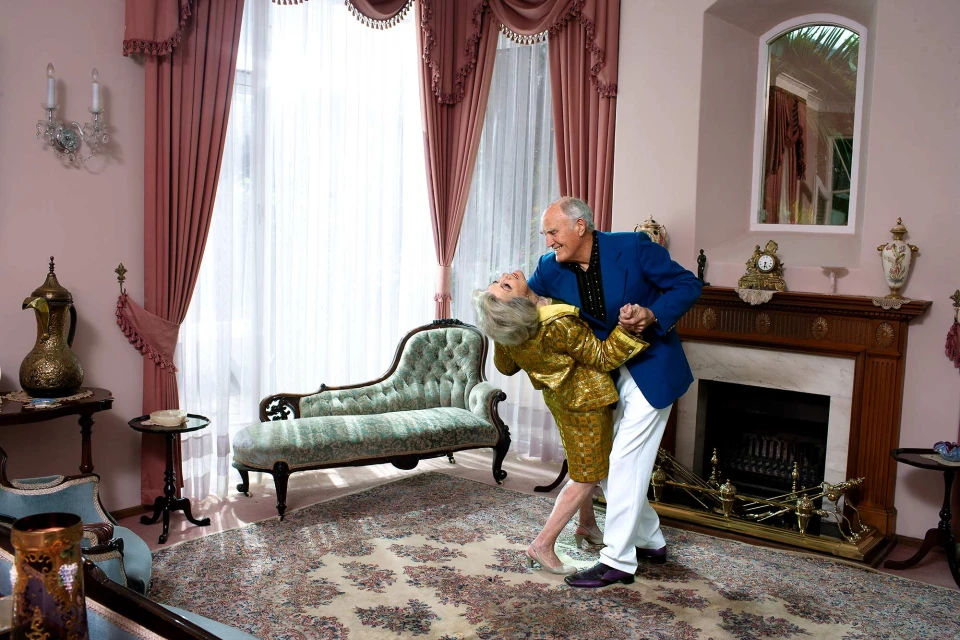 Senior couple in formal wear dances together in a grand, ornate living room with vintage furniture, pink walls, and floral patterned carpet.
