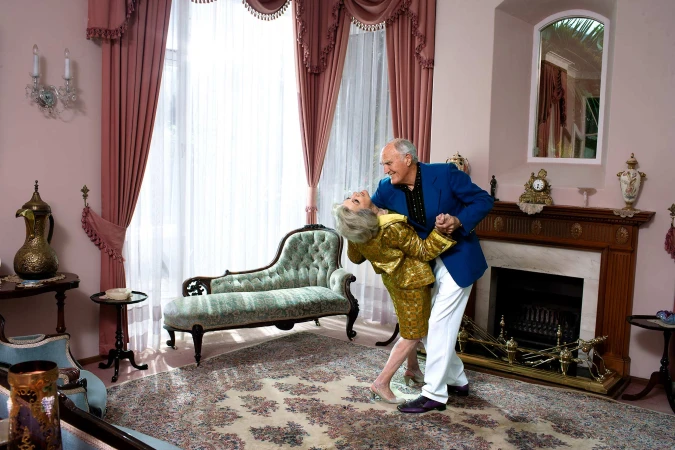 Senior couple in formal wear dances together in a grand, ornate living room with vintage furniture, pink walls, and floral patterned carpet.