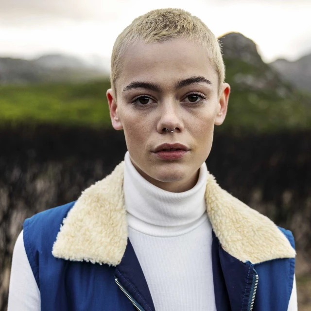 Portrait of a young adult woman with a shaved head, wearing a blue vest and turtleneck, against an outdoor background with mountains.