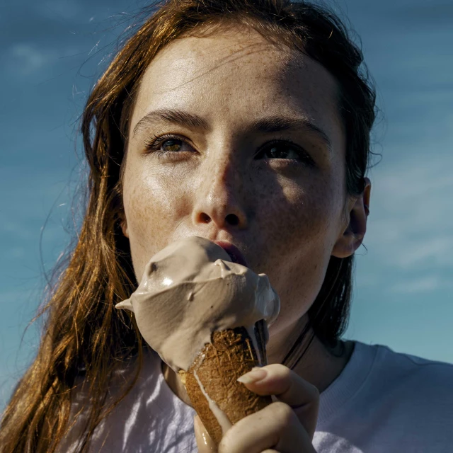 Close-up of a young woman with freckles eating chocolate ice cream cone outside on a sunny day with a blue sky background.