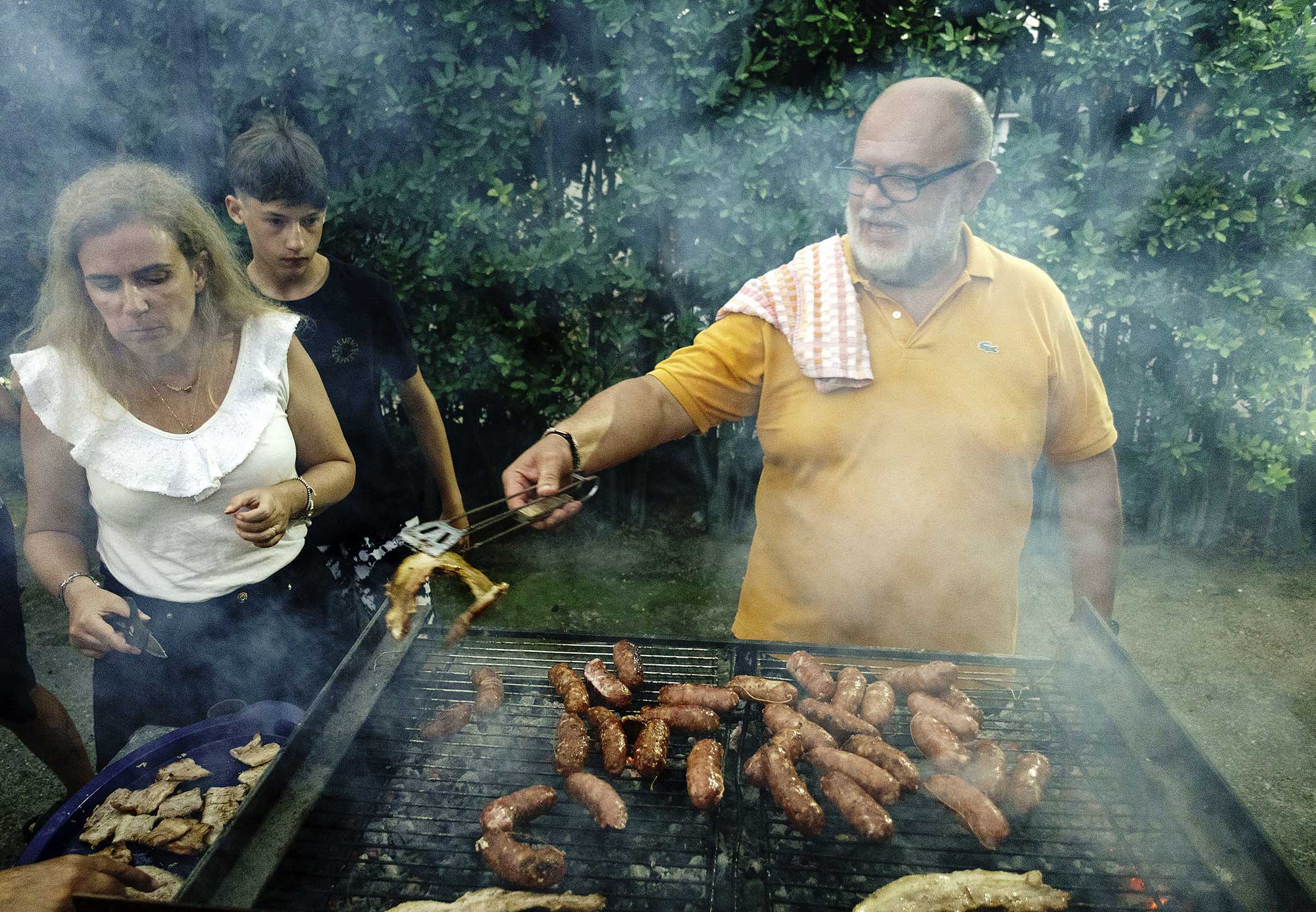 Three people at an outdoor barbecue. An adult man tends to sausages and chicken on a grill, while an adult woman and a teen watch.