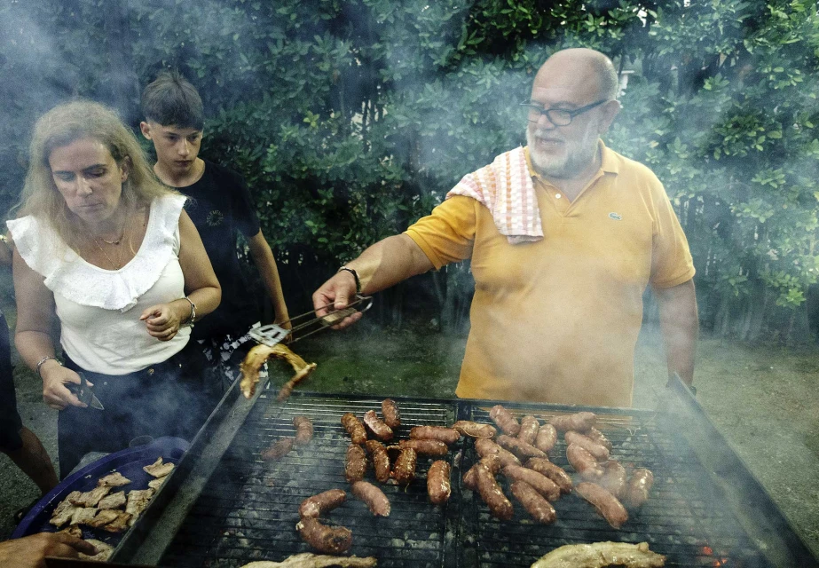 Three people at an outdoor barbecue. An adult man tends to sausages and chicken on a grill, while an adult woman and a teen watch.