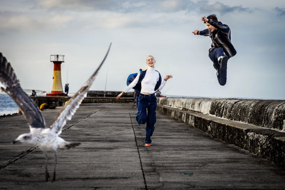 Two people in motion near a lighthouse and body of water, one woman running, one man mid-air, with a seagull in foreground.