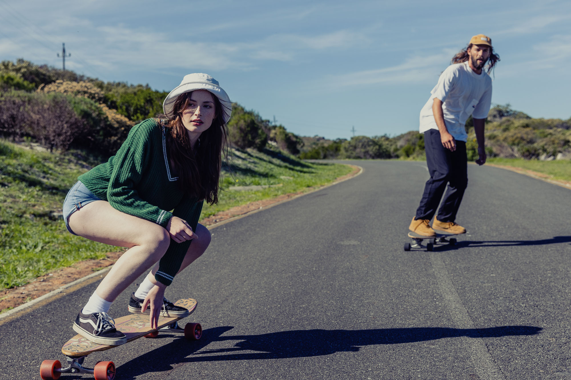 Two people skateboarding on an asphalt road under a bright, sunny sky with green vegetation in the background and one woman in the foreground.