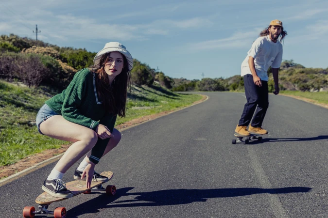 Two people skateboarding on an asphalt road under a bright, sunny sky with green vegetation in the background and one woman in the foreground.