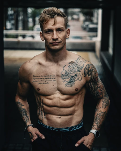 A muscular man stands shirtless, showcasing tattoos and fitness, in a dimly lit area. The lighting highlights his physique and definition.