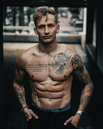 A muscular man stands shirtless, showcasing tattoos and fitness, in a dimly lit area. The lighting highlights his physique and definition.