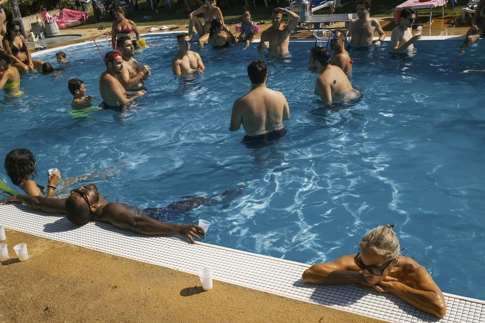 Large group of people in outdoor swimming pool relaxing in the water on a sunny day, including adults and children.