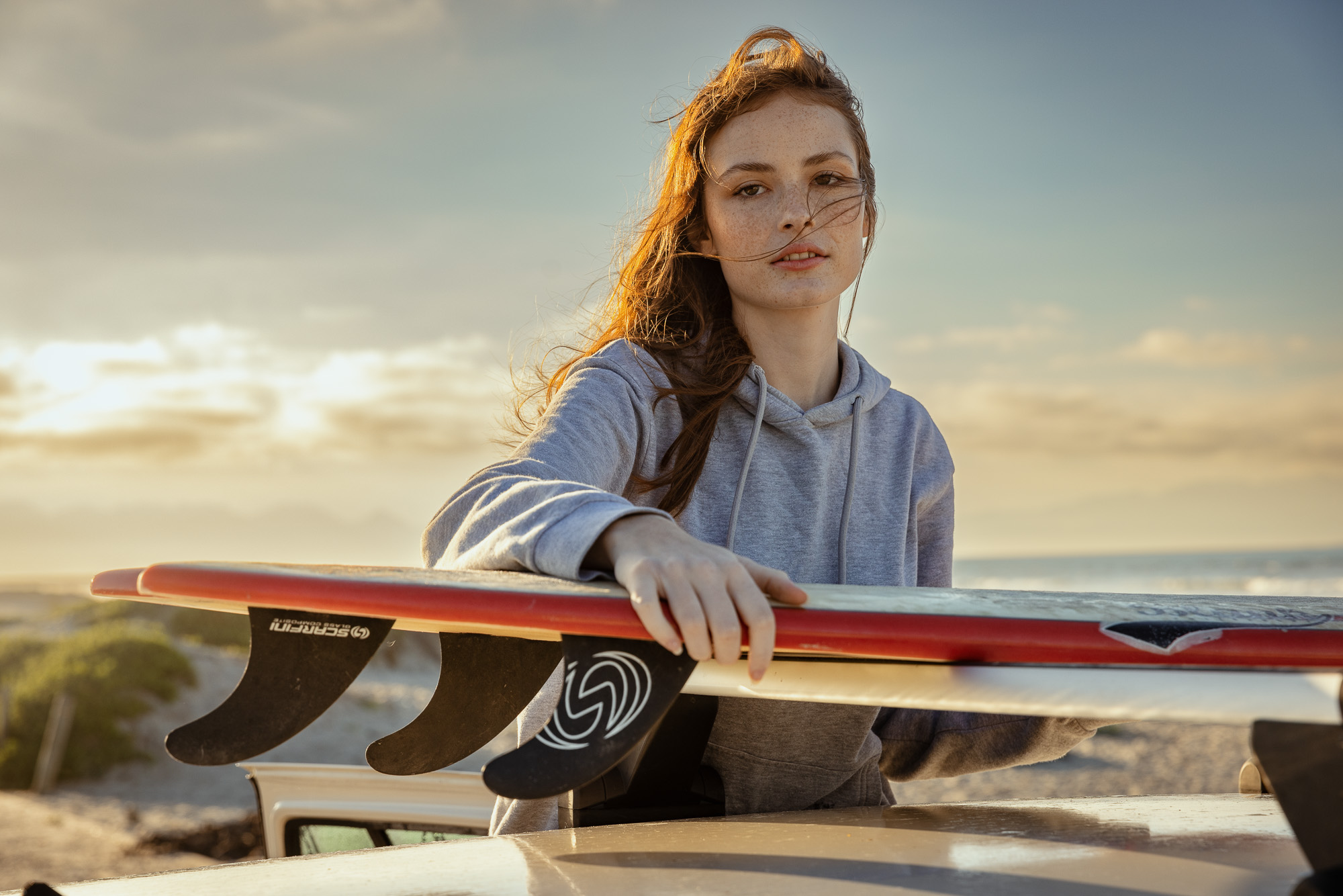 Portrait of a red-haired woman in a grey hoodie, standing behind surfboards on a sandy beach with a sunset-lit sky in the background.