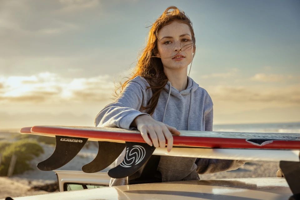 Portrait of a red-haired woman in a grey hoodie, standing behind surfboards on a sandy beach with a sunset-lit sky in the background.