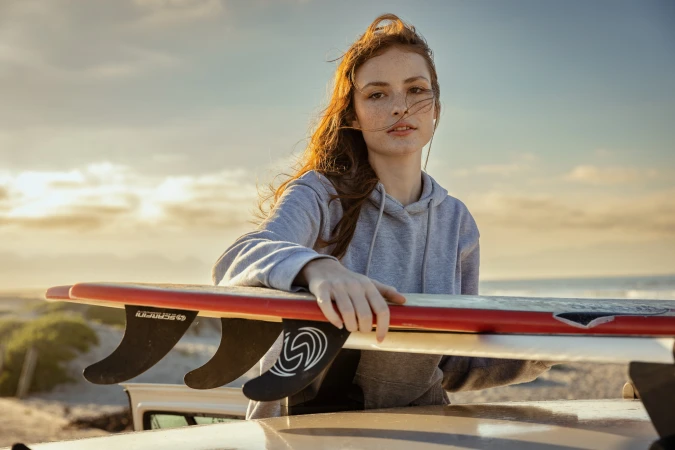 Portrait of a red-haired woman in a grey hoodie, standing behind surfboards on a sandy beach with a sunset-lit sky in the background.